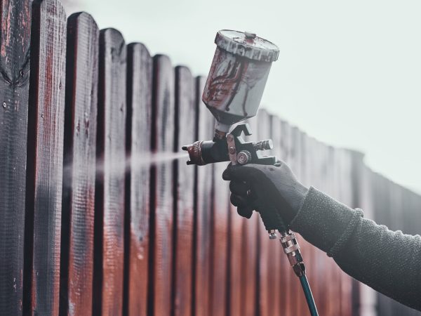 Process of old fence renovation by worker in protective gloves. He is usin airbrush, applying paint.