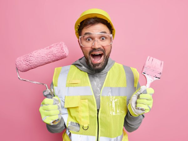 Positive male professional handyman holds painting roller and brush looks gladfully at camera happy to finish doing repairing dressed in uniform isolated over pink background. Maintenance work