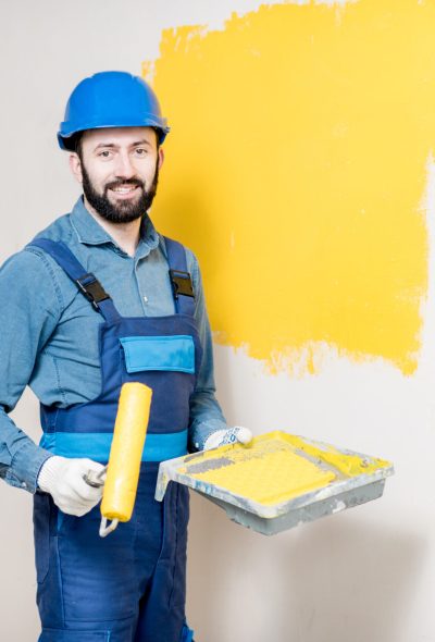 Portrait of a handsome painter in working uniform painting wall with yellow paint